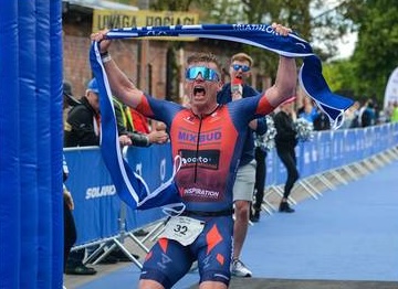 Triathlonista świętuje linię mety z flagą narodową i banerem zawodów - plan treningowy triathlonowy IM Inspiration